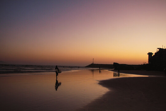 surfer walks along the beach in Imsouane, Morocco, carrying his board at sunset under purple and golden evening light