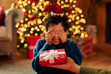 Joyful child boy covering his eyes with hands, receiving gift box held by an adult, beautifully decorated Christmas tree on the background