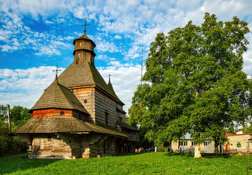 Church of the Exaltation of the Holy Cross, a historic wooden Ukrainian Greek Catholic church, stands on a grassy hill with a lush green tree under a bright blue sky with white clouds