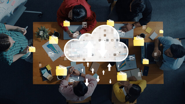 Diverse team collaborates at a wooden table with digital cloud icons representing file sharing and uploads, showcasing modern technology and teamwork in an office setting. Trope - Powered by Adobe