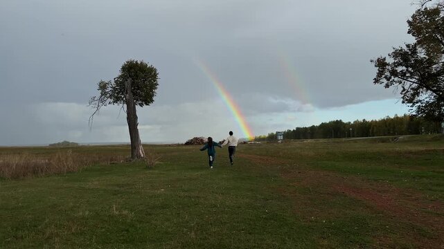 Romantic adventure in open field as man and woman race side by side towards rainbow, solitary tree and sunset colors highlight peaceful joy.