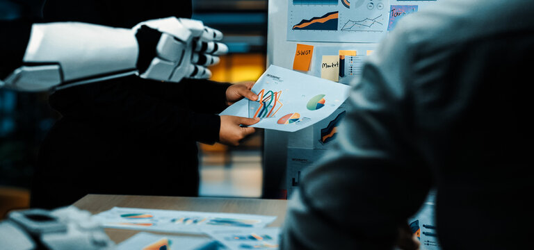A futuristic business meeting highlights collaboration between humans and robots, focusing on charts and graphs to enhance decision-making in a modern office environment.Noogenesis - Powered by Adobe