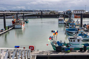 a vibrant harbor scene in Royan, with colorful fishing boats docked in calm water under a partially cloudy sky. Buildings and infrastructure line the distant shore.