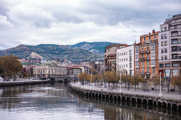 wide view of the Bilbao riverfront, with colorful buildings reflected in the calm water. The city rises up a wooded hillside under a dramatic, cloudy sky, showcasing urban and natural elements.