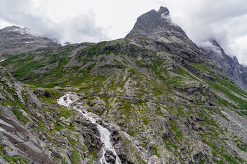 Obraz premium Remote houses at de foot of the Bispen mountain towers above the lush rocky landscape, with the Stigfossen waterfall cascading down its rocky slopes near the scenic Trollstigen road in Norway