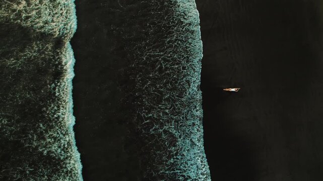 Top down of woman alone sunbathing on dark sand beach with breaking ocean waves