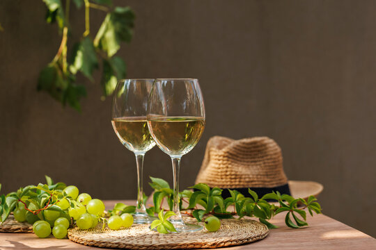 Two white wine glasses with green grapes and hat on rustic outdoor table with soft sunlight..