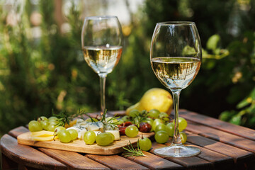 Naklejka premium Two wine glasses with white wine and cheese board served with grapes and pear on wooden table outdoors