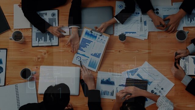 Overhead view of a collaborative business meeting. Professionals analyze charts and data on a wooden table, engaging in strategic discussions to drive success. Raster