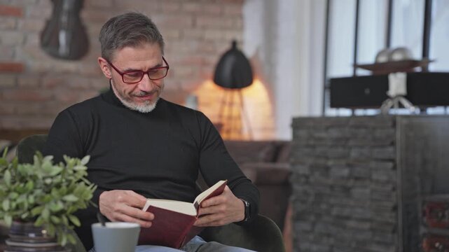 Smiling middle aged man sitting in living room reading book. Happy male wearing turtleneck pullover and eyeglasses. Smart looking older guy with grey hair enjoying leisure and free time at home.