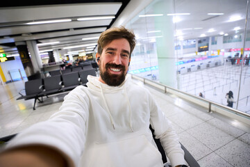 Bearded millennial man taking a selfie at a bright airport terminal before boarding. Casual hoodie, backpack and earbuds, smiling while waiting, with modern architecture behind.