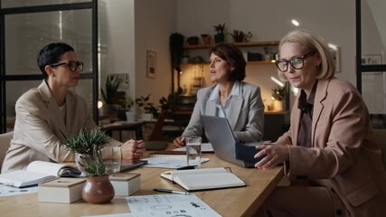 Three professional middle-aged Caucasian women in formal outfits collaboratively working in modern office workspace