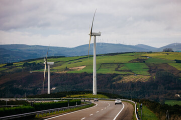 Naklejka premium Wind turbines stand tall on rolling green hills beside a winding road, showcasing renewable energy and sustainable technology in a serene landscape