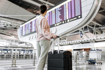 Female traveler with suitcase stands in airport terminal, gazing at flight information display board, surrounded by modern architecture and bustling atmosphere, ready for journey © wedmoments.stock