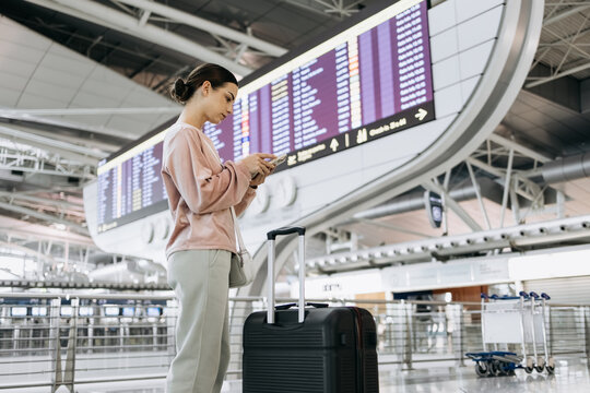 Young woman with dark hair, wearing a pink sweater, is checking her phone while standing beside a suitcase in a modern airport terminal with flight information display