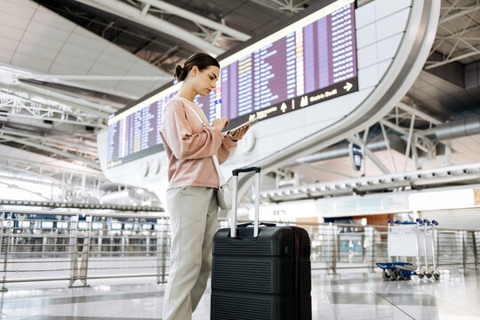 Young woman with dark hair stands at airport terminal, checking her smartphone while waiting for flight, with departure board and luggage in the background, capturing travel anticipation