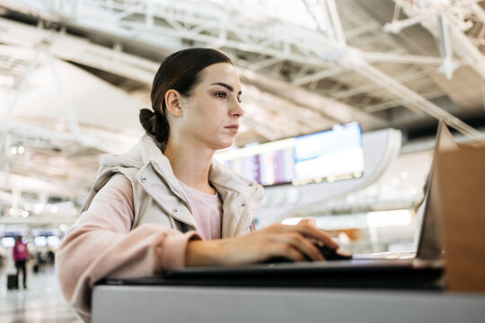 Young woman with dark hair, focused on laptop in modern airport terminal, surrounded by travelers, showcasing digital connectivity and travel experience