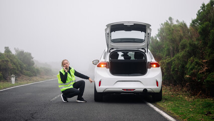Obraz premium A woman wearing a reflective vest kneels next to a broken down car on a foggy road, demonstrating emergency repair and assistance.