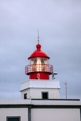 Red lighthouse with a glass lantern, standing tall against a cloudy sky, symbolizing guidance and safety for maritime navigation in coastal regions