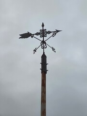 Ornamental weather vane standing tall against moody clouds, its dark silhouette and rustic texture creating a timeless countryside atmosphere.