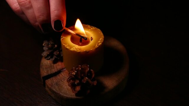 Woman hand lights a match the bees wax candle on wooden candlestick with decor Christmas cones on dark black background. Person lighting the decoration handmade single candle for home atmosphere, mood