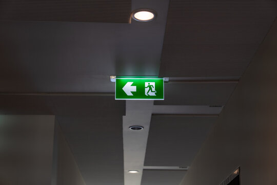A green fire escape sign hangs from the ceiling, showing an exit sign green light box with a running man and an arrow pointing right for safety and evacuation.