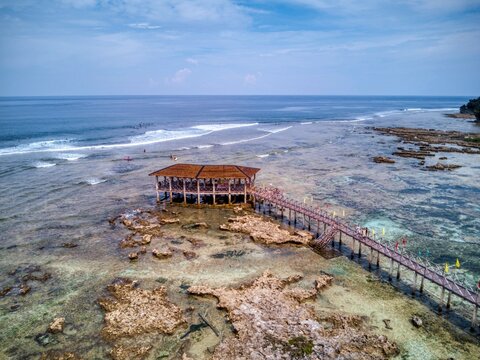 Drone view of boardwalk pavilion over rocky tidal flats with surfers beyond