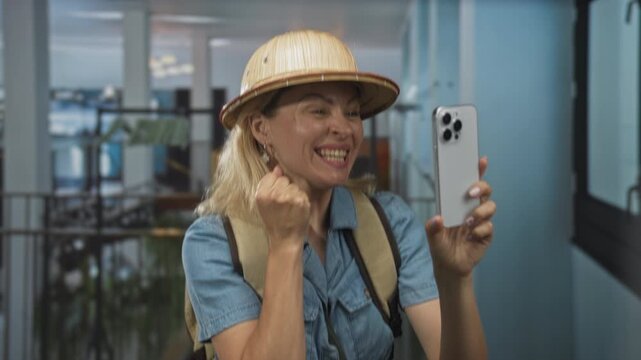 Young blonde woman holds smartphone with two hands and clenches fist in airport terminal; excitement.