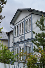A traditional wooden house with ornate trim and large windows stands under a cloudy sky, surrounded by greenery and a white picket fence in Jurmala, Latvia