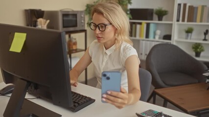 Blonde woman with glasses holds smartphone at white computer desk in bright office; concentration efficiency.
