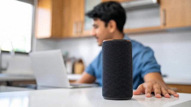 A smart speaker sits on a white surface with a laptop and person in the background