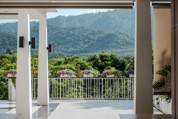 Fototapeta premium A bright terrace overlooking green hills features rows of colorful flowers along the railing, with clean white pillars framing the open space and highlighting the mountain scenery.