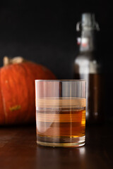 A vintage rocks glass partially filled with brown alcohol, bottle and orange pumpkin in background
