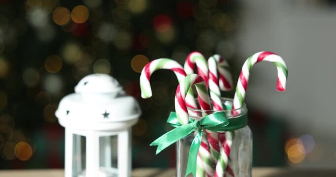 Merry Christmas and Happy New Year. Jar with tasty candy canes and lantern on light wooden table against blurred lights, closeup. Camera moving