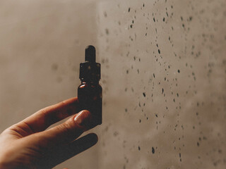 Hand holding a small brown essential oil bottle with a dropper. A hand gently holds a small bottle of essential oil, with water droplets on the glass behind it. Woman in a bathroom. Beauty routine.