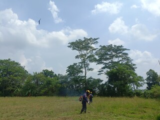 workers whose job is to cut grass in the open field area for oil and gas extraction, working under the bright blue sky and green nature, the engine also smokes but it is safe if workers wear mask