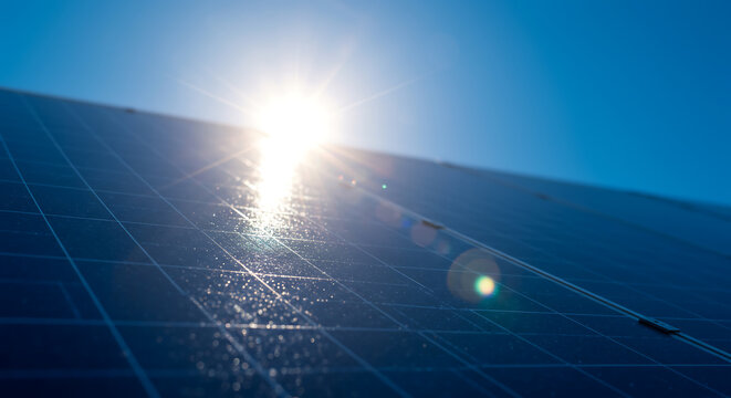 Bright sun shines directly onto a clean solar panel, creating a lens flare and reflections against a clear blue sky