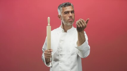 Man chef with grey hair in white jacket holds wooden rolling pin, gestures beckoning with bare hand against red studio backdrop; confidence.