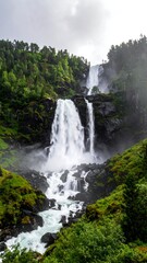 Fototapeta premium A majestic waterfall cascades down a rocky cliff, framed by lush green forest and a dramatic, overcast sky. Water flows strongly