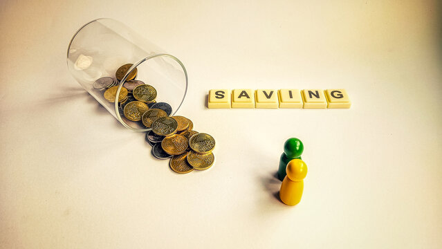 Spilled coins from a glass jar with wooden figures and the word SAVING, symbolizing financial planning, budgeting, and money management for the future.