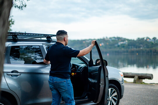 A man in his thirties stands beside his car, taking in the serene lakeside view during a trip
