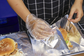 Chef hand in glove carefully wrapping fresh burger in aluminum foil for takeaway. focused cook preparing fast food with hygiene in professional kitchen for service