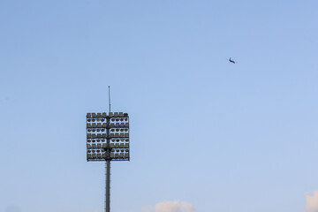 Airplane flying high in blue sky above tall stadium floodlight. This peaceful outdoor scene suggests travel, freedom, and wide open expanse of atmosphere