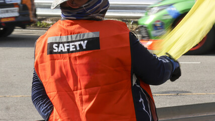 Professional race marshal from behind wearing an orange safety vest and white helmet. man holding yellow flag watches over track with focused responsibility during motorsport event