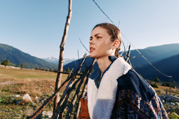 Woman portrait in mountains countryside near rustic fence, wearing warm shearling jacket and patterned sweater, looking thoughtful at sunny landscape during outdoor travel.