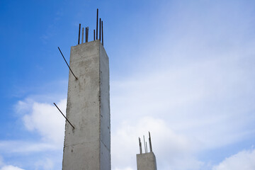 Towering raw concrete pillars with visible rebar extend upwards into a bright blue sky dotted with scattered white clouds, symbolizing ongoing construction and development