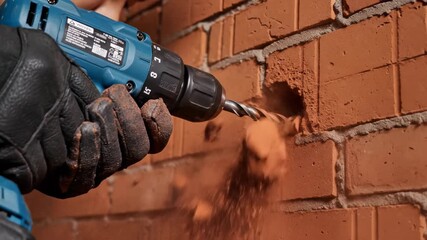 Hand in black gloves using a power drill on a red brick wall, creating dust, concept for home renovation, construction and building maintenance and repair services