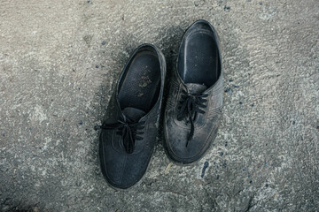 pair of well-worn, dusty, black canvas shoes (likely a sneaker or plimsoll style) resting on a rough, textured grey concrete or cement surface. The image emphasizes the simple, utilitarian nature