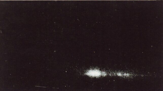 A dark image featuring a faint cluster of light, possibly depicting celestial phenomena or astrophotography.