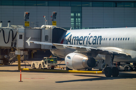 Calgary, Alberta, Canada. Nov 22, 2025. An American Airlines Airbus A320 is serviced by ground crew and jet bridge at a sunlit airport terminal gate, ready for passenger boarding.